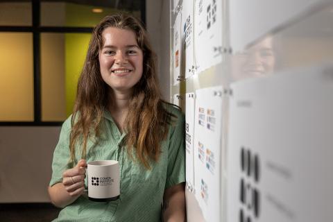 A female student, Julia Baer, stands in front of a white board holding a mug with the Cohen Institute for Holocaust & Genocide Studies logo on it